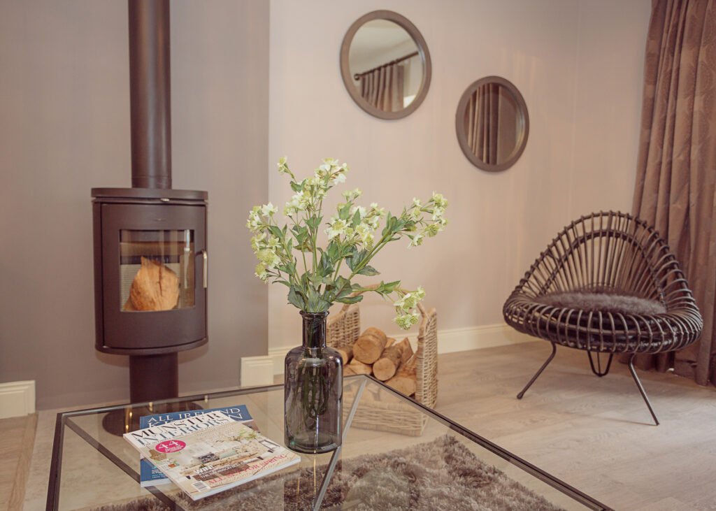 Living room detail with glass coffee table, fresh flowers, wood-burning stove and rattan lounge chair – Innishannon Showhouse by AK Interior, Cork Ireland.