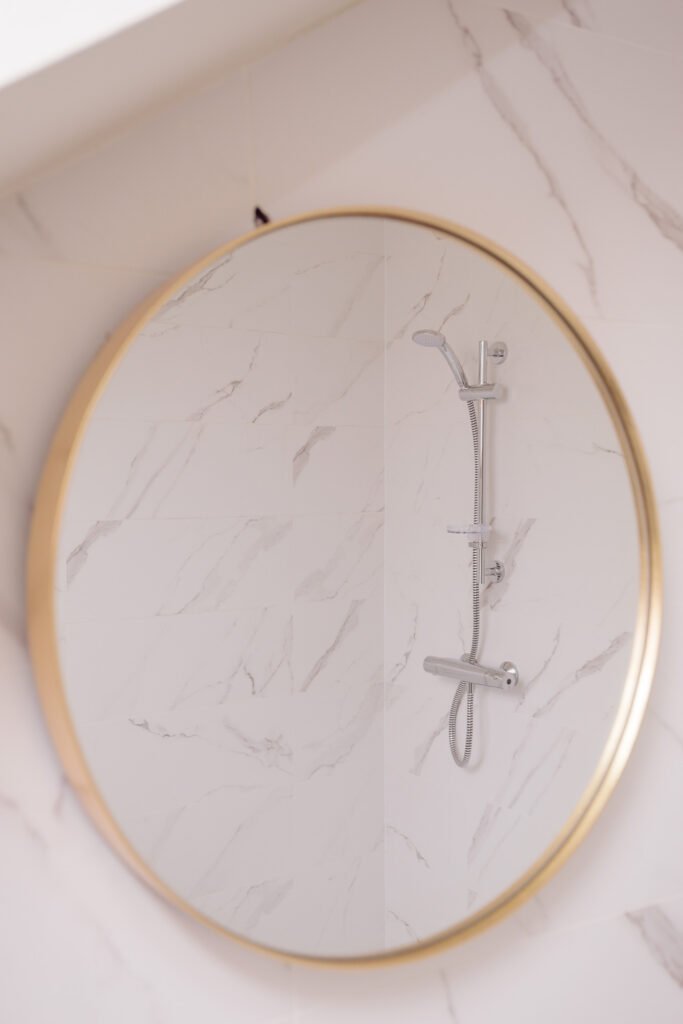 Round mirror reflection showing modern marble-tiled shower with chrome fixtures – Innishannon Showhouse by AK Interior, Cork Ireland.