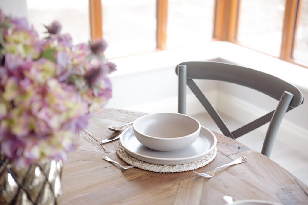 Close-up of table setting with stacked ceramic dishes, cutlery and a floral arrangement on a wooden dining table – Innishannon Showhouse by AK Interior, Cork, Ireland
