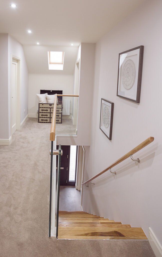 Bright hallway with glass balustrade staircase, framed wall art and skylight – Innishannon Showhouse by AK Interior, Cork Ireland.