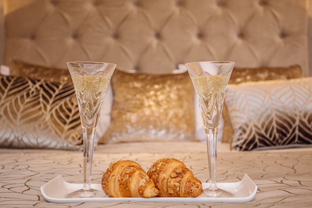 Close-up of champagne flutes and croissants on patterned bedspread with gold cushions behind. Rockport Residential project, AK Interior, Cork.