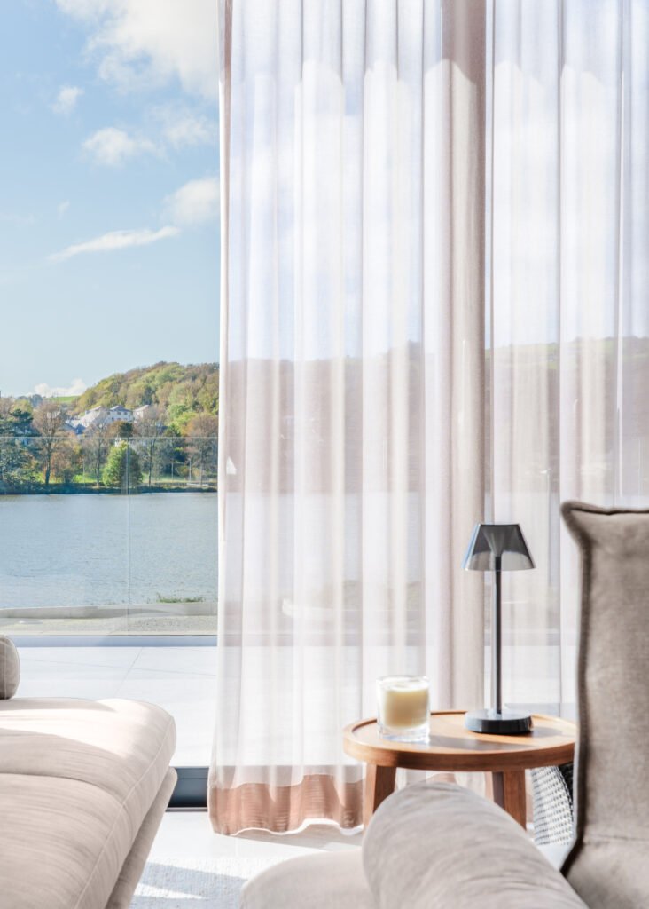 Waterfront view through sheer curtains with beige armchair and round side table – West Cork Waterfront, residential project by AK Interior, Cork, Ireland.
