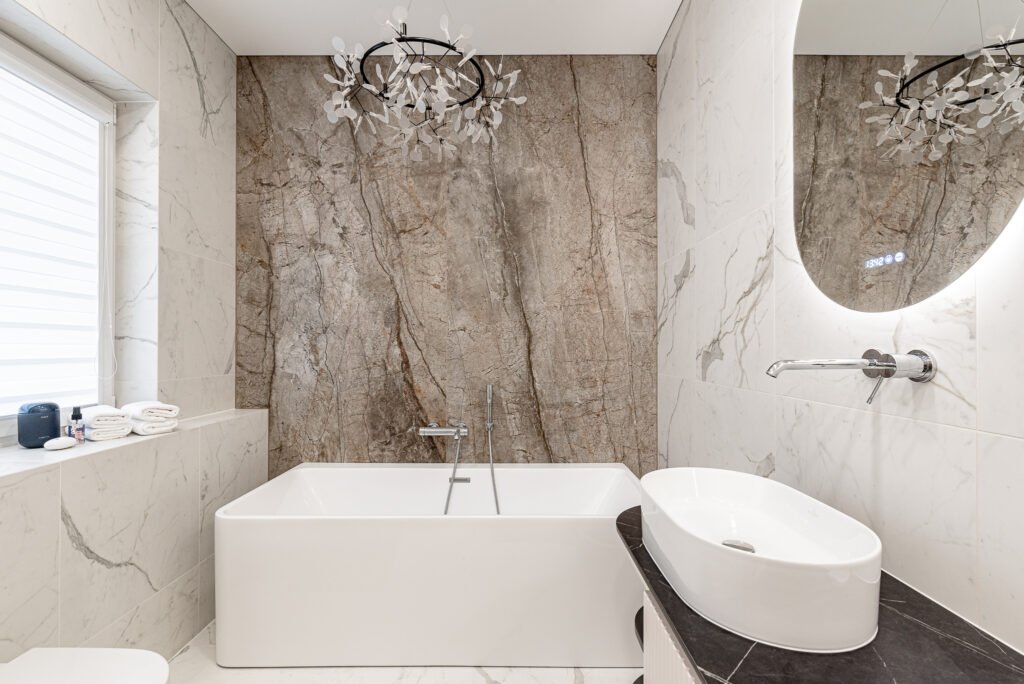 Modern bathroom with a stone-textured feature wall, deep bathtub and curved mirror – AK Interior, Monkstown Full House Design